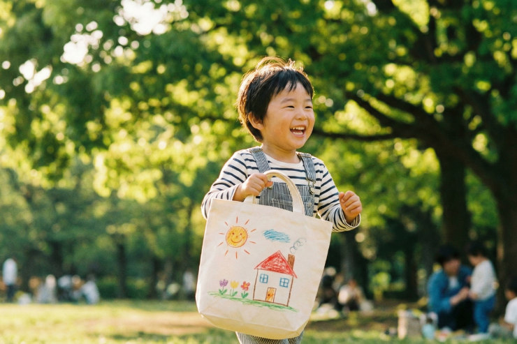 自分の絵がプリントされたトートバッグを肩にかけて公園でポーズをとる日本人の子供